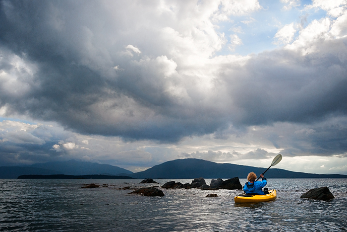 Blond woman paddles a kayak around rocks in the bay, Indian Cove, Juneau, Alaska